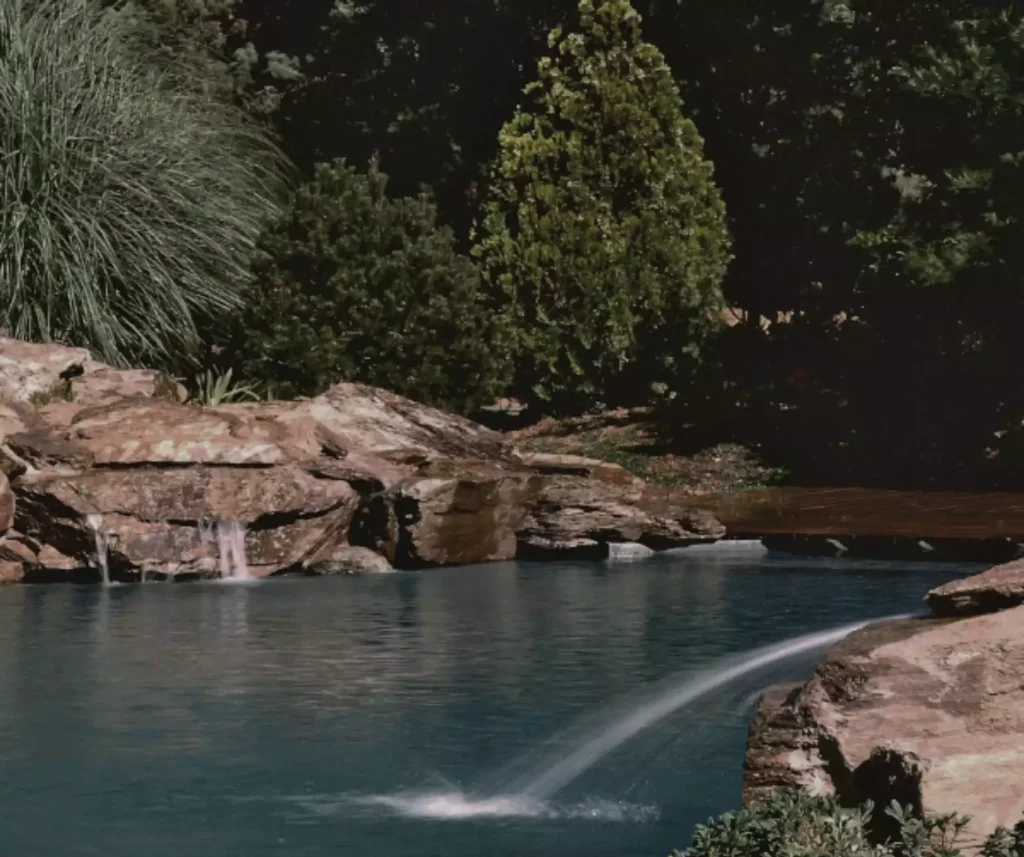 A natural rock pool water feature with cascading waterfalls and a curved water jet, seamlessly integrated into a lush, landscaped setting.