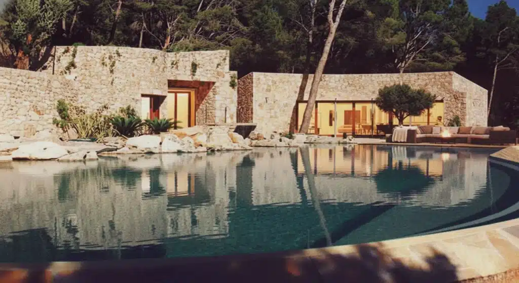 Curved infinity pool integrated into a sloping hillside with traditional dry-stone walls and pine trees at sunset in Formentor, Mallorca – Morana Water Design